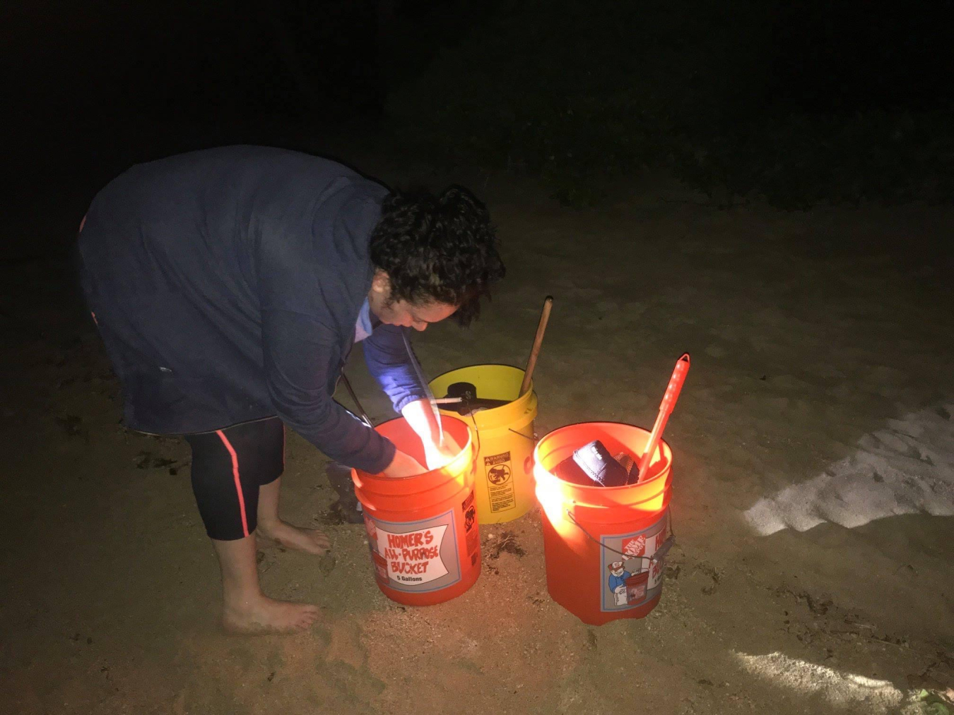 Nina bending over three buckets on a beach at night.