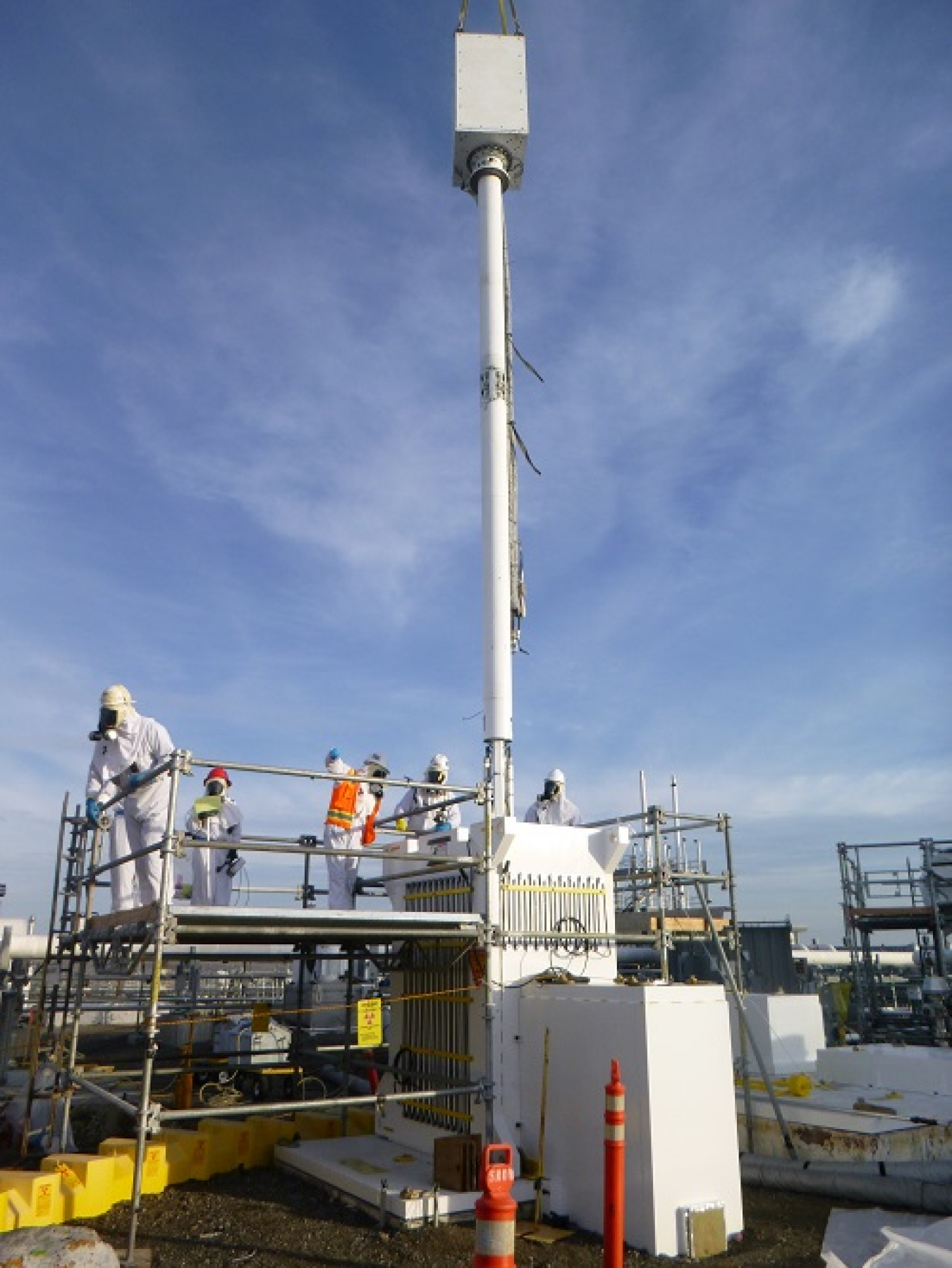 A tall white pole coming out of the ground, workers stand around on a platform beside it and perform work