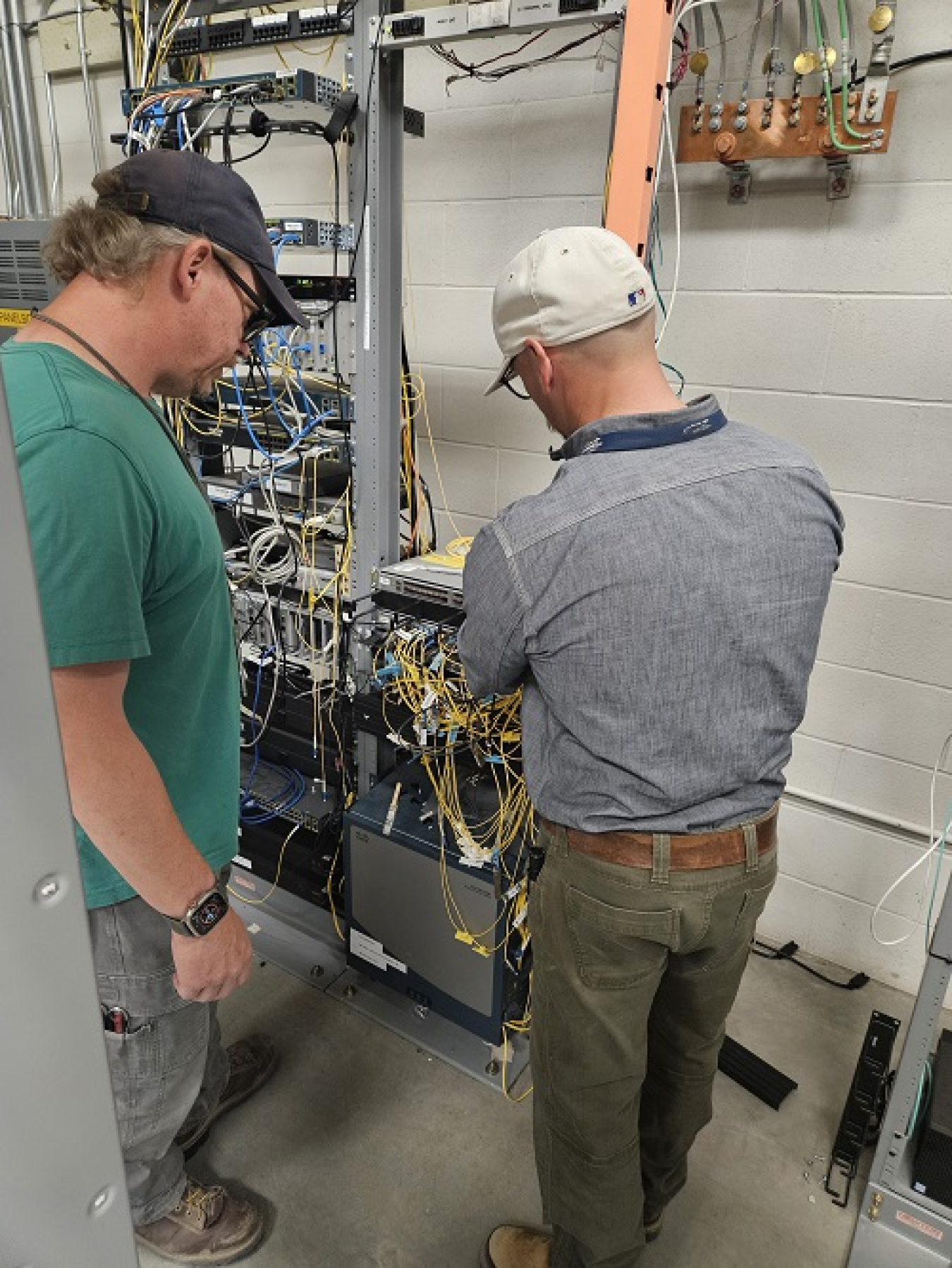 Two men stand in front of machines with lots of blue and yellow wires coming out,