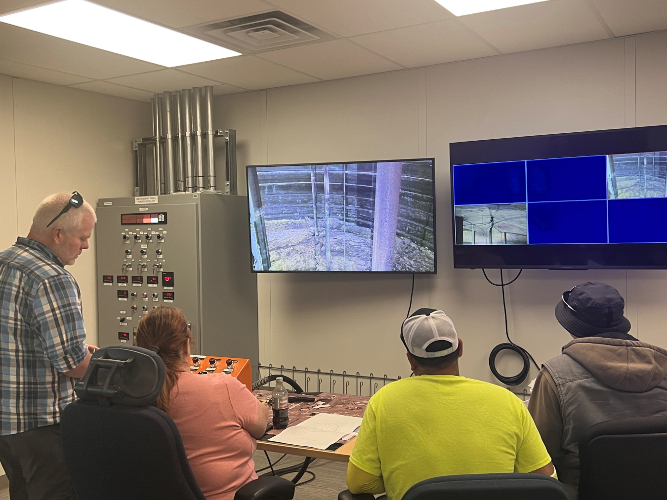 A group of people sit in a control room and watch two screens mounted on the wall in front of them