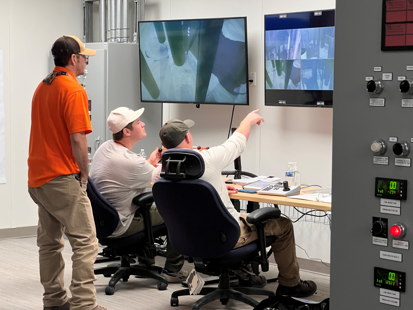 Three employees in a control room look up at a monitor, one points to the monitor