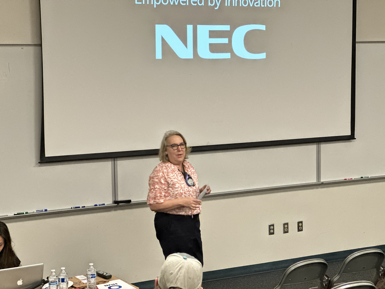 A woman stands at the front of a lecture room, there is a powerpoint screen behind her that says "NEC"