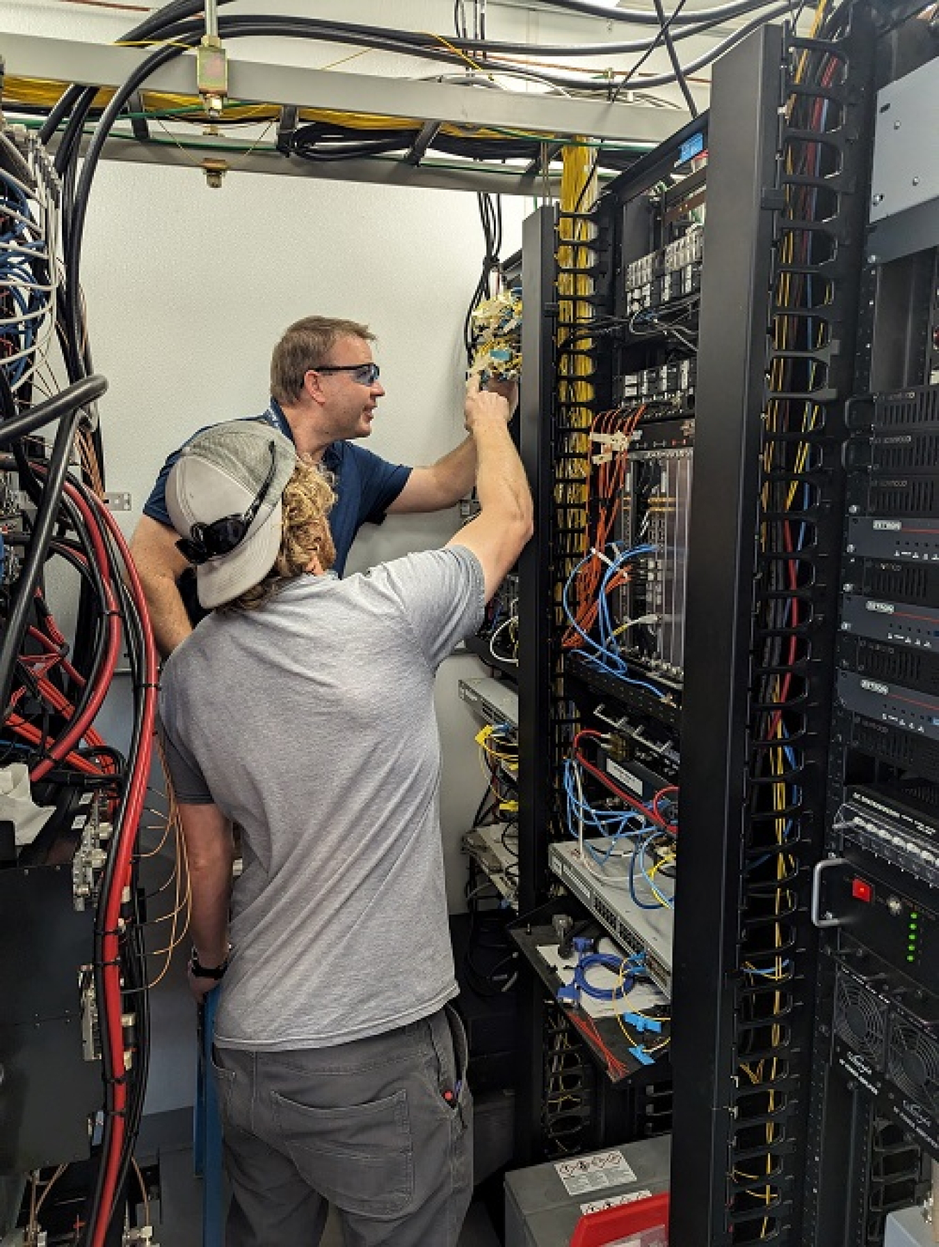 Two people work in a facility building on a wall with machines and lots of colorful wires coming out of them