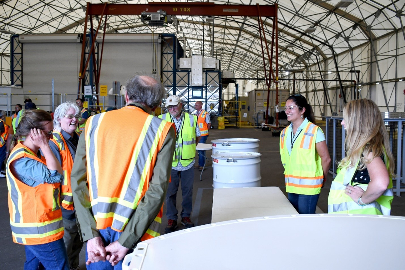 People stand inside a large building, they wear safety gear and stand beside white tanks