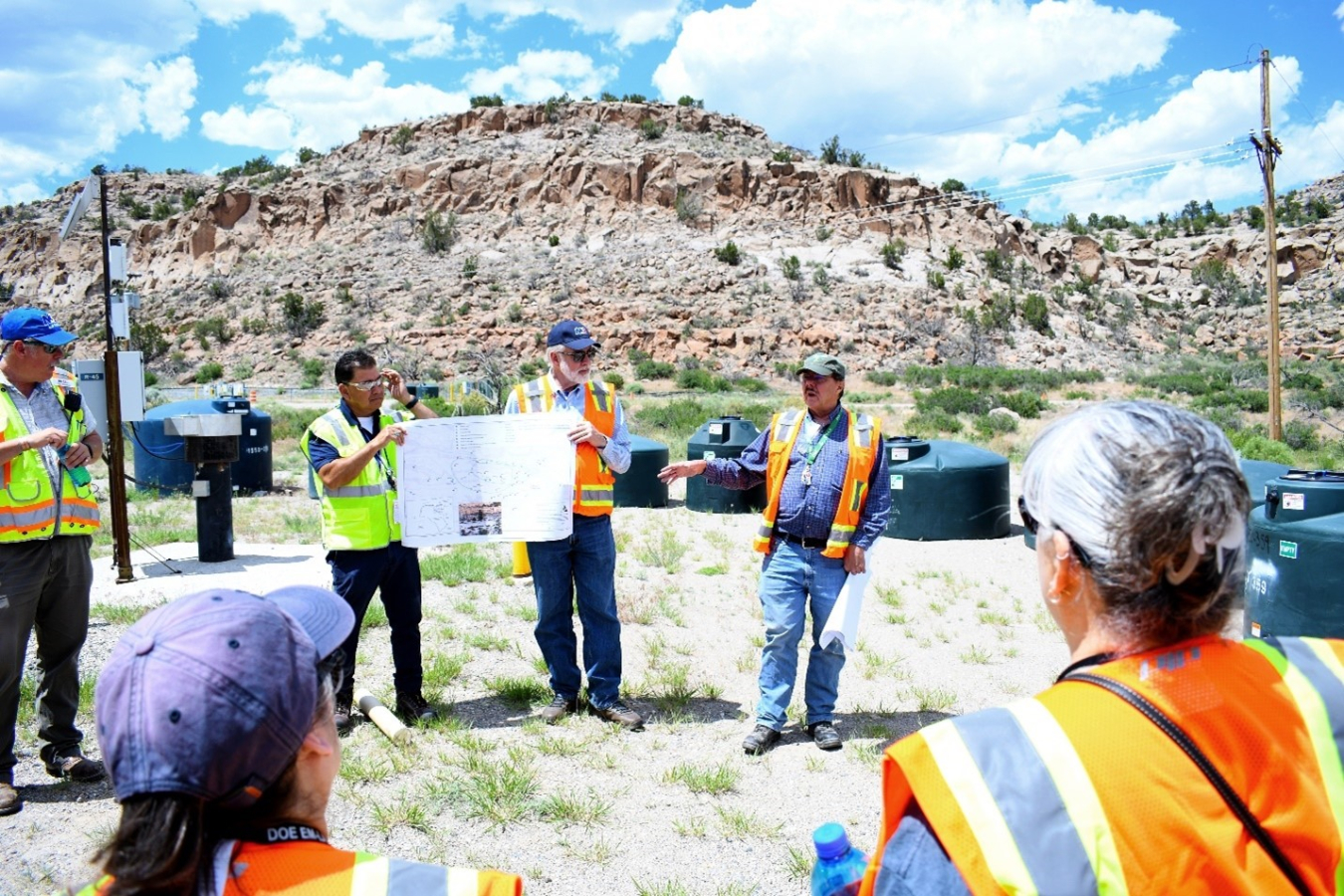 Individuals stand outside with orange and yellow safety vests on, two individuals hold up a poster while the others look at the poster
