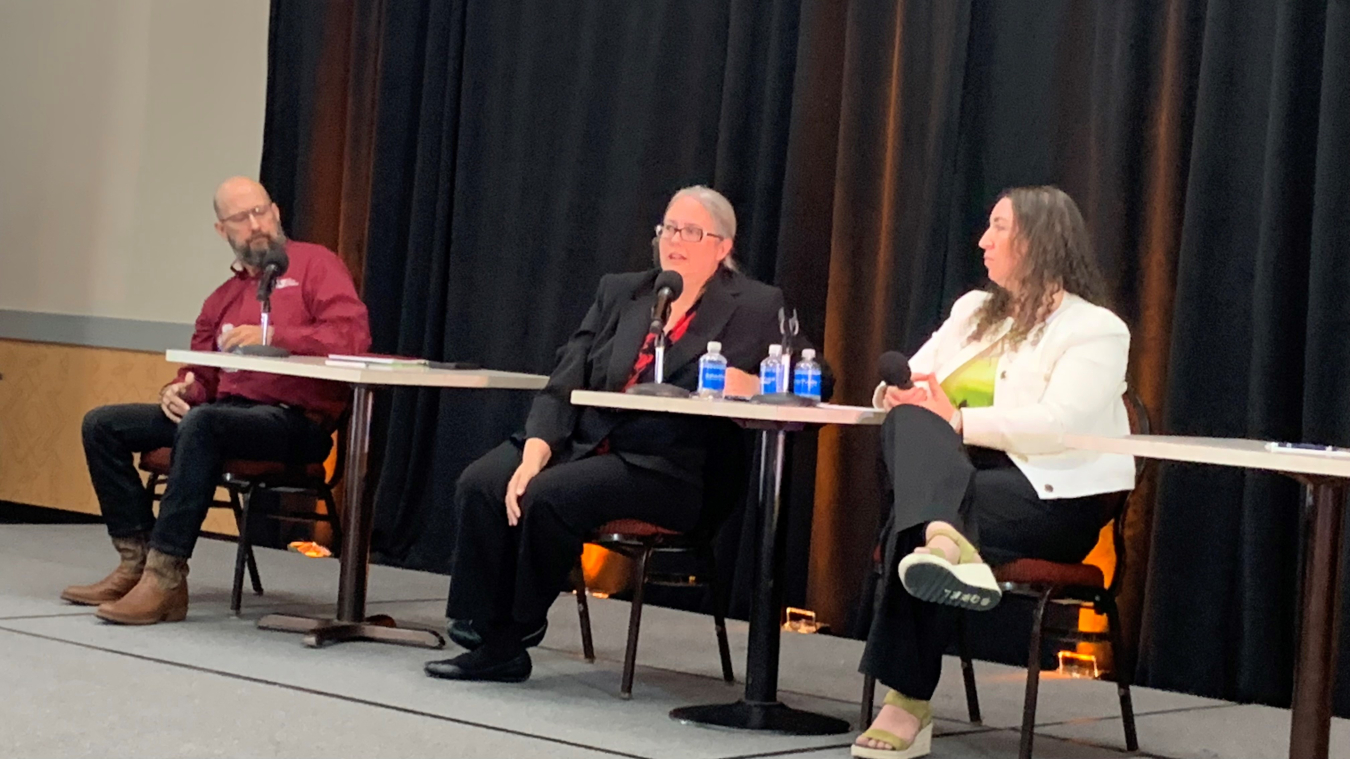 A group of people sit at a panel table at the front of a room and look at the audience