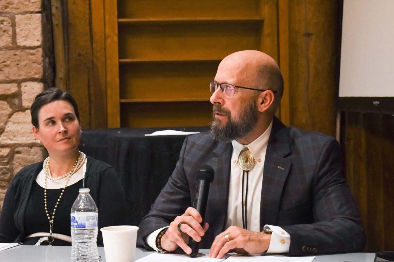 A bald man sits at a table and speaks into a microphone, a woman sits beside him and looks at him while he speaks