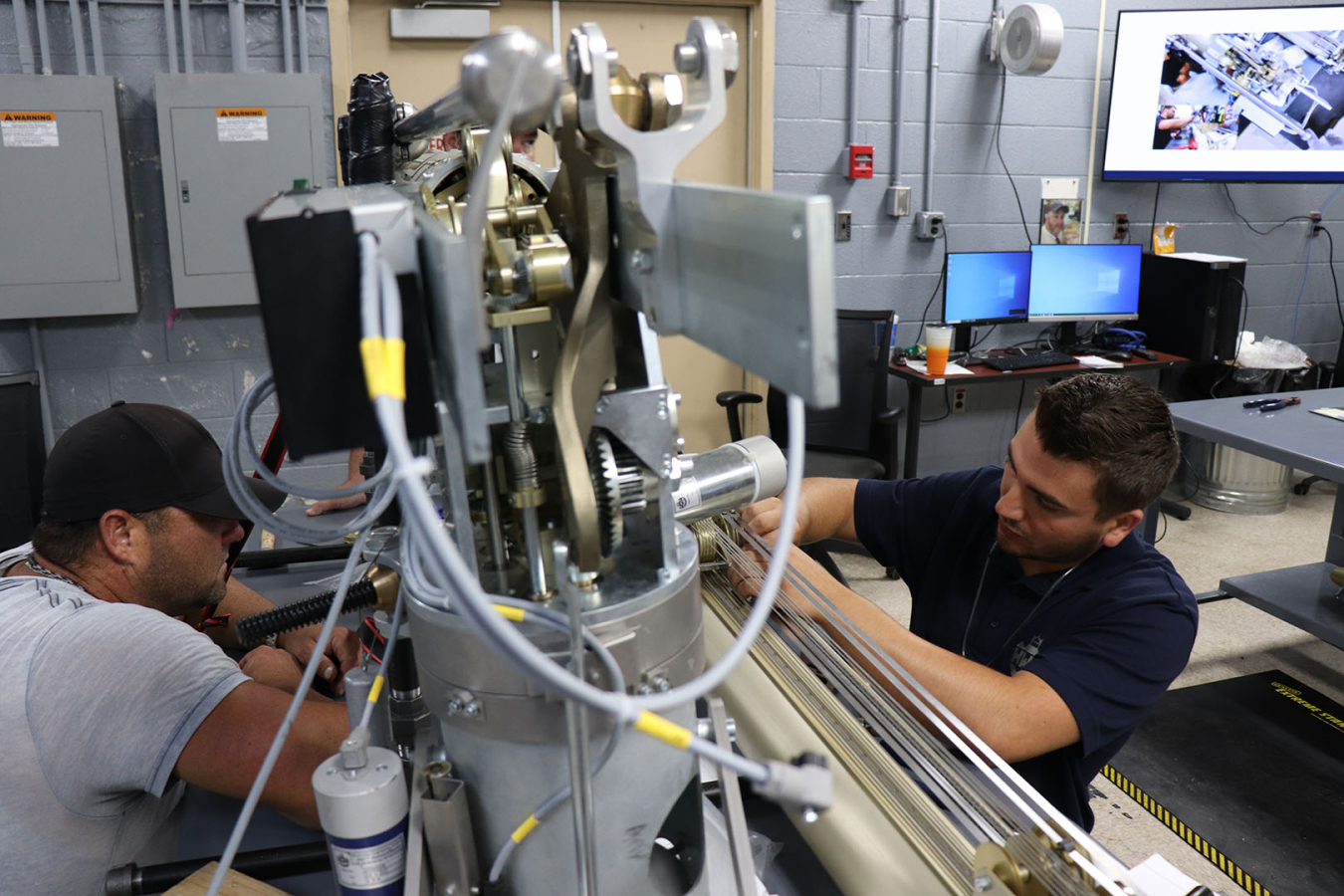 Two men work on either side of a gray machine inside a facility building