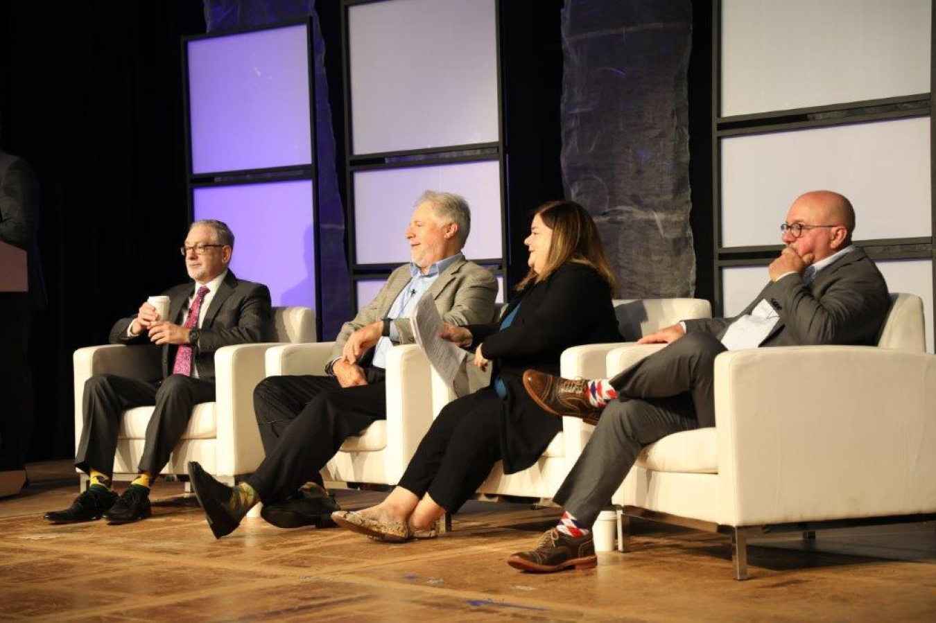 Four professional sit on while chairs on a stage and talk and face the audience