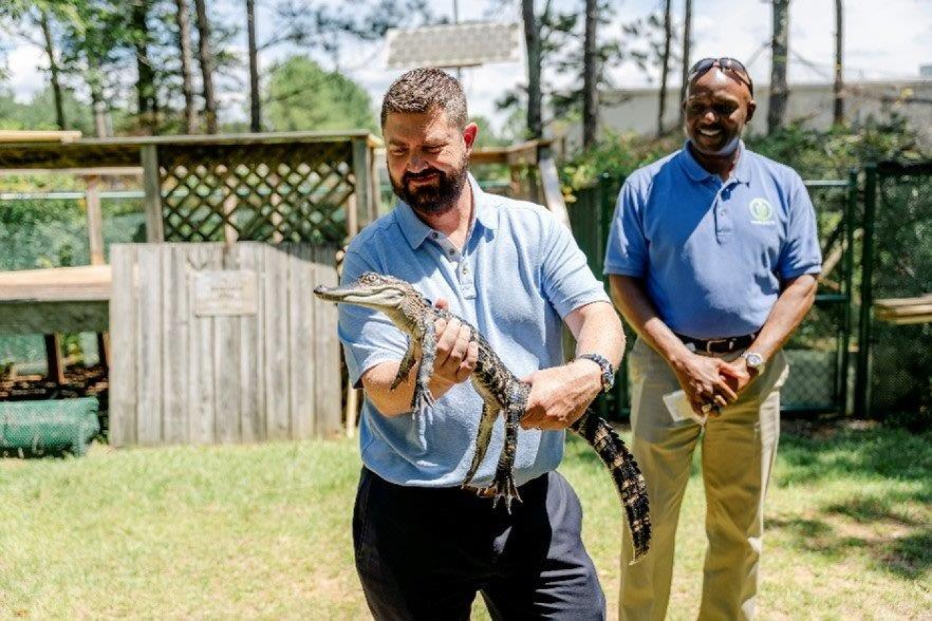 A person holds a juvenile alligator with a person standing behind.
