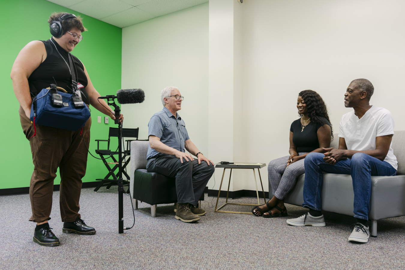Four people in a room, two sit on a couch and one on a chair, the other stands up and uses a camera to record the others