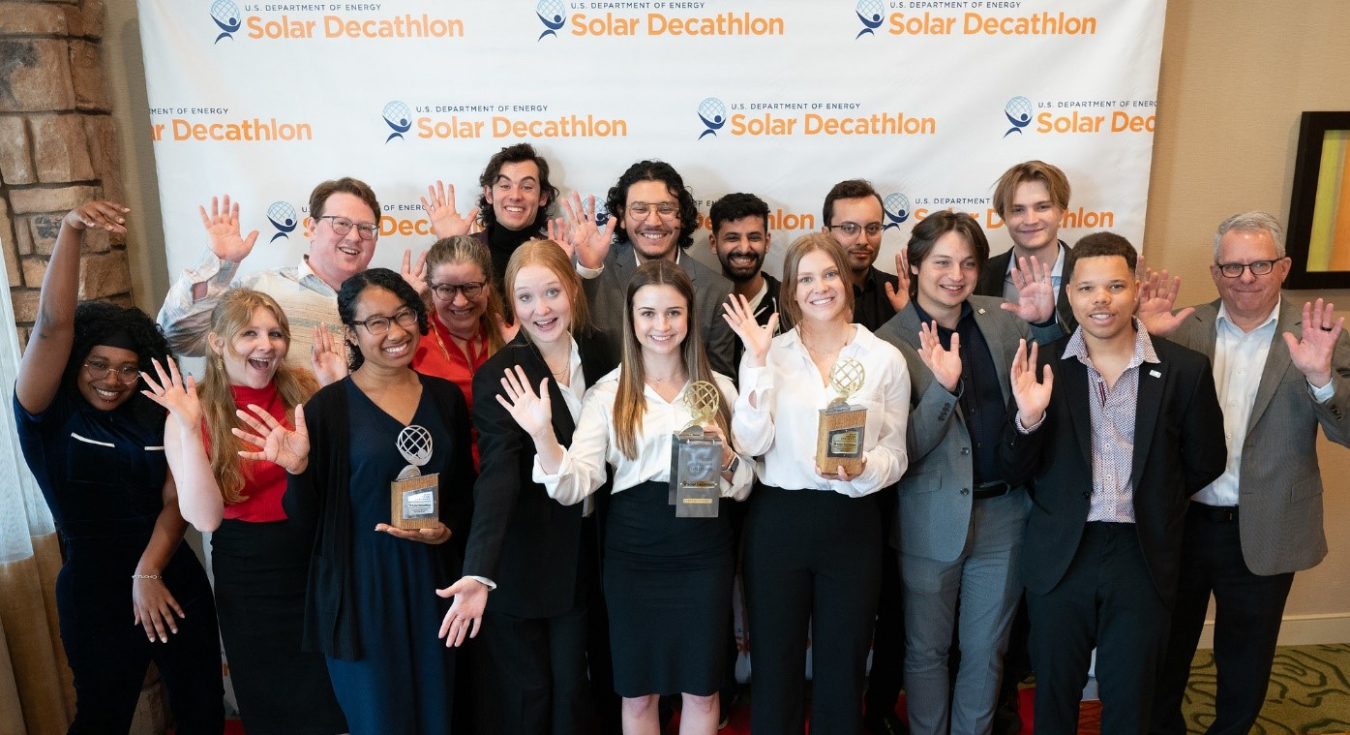 Group of student competitors standing and facing the camera, waving their hands and holding trophies.