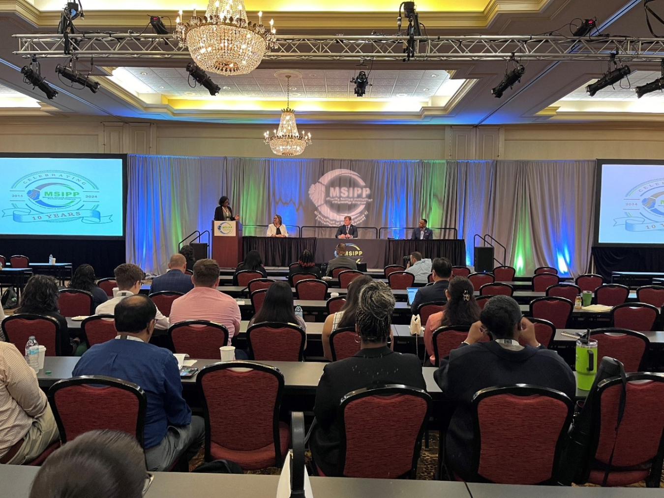 A large ball room with people sitting at tables facing the stage while a panel takes place on the stage