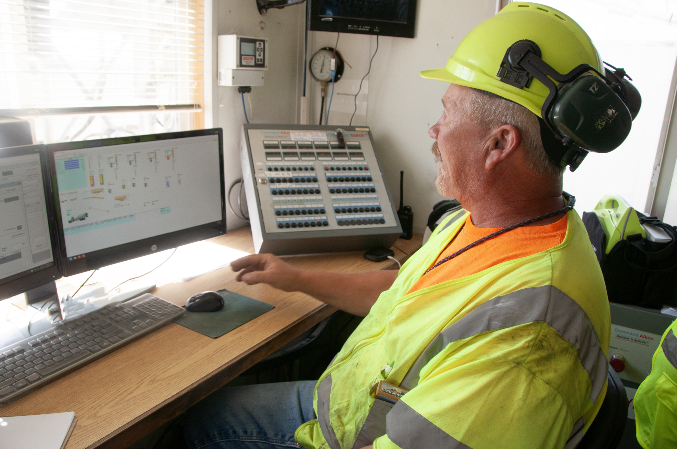 A man wearing a yellow and orange safety vest and hard hat sits at a desk and uses a computer