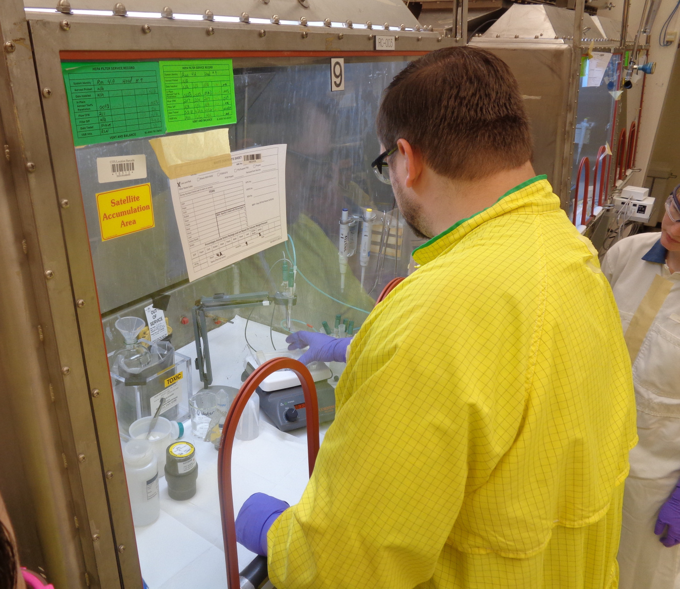 A man in a yellow safety jacket stands with his back towards the camera and works inside a glass covered area