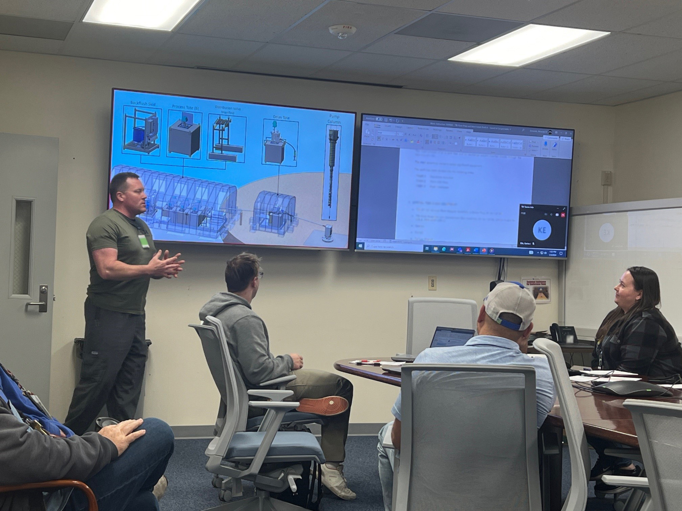 A group of people sit at a table in a conference room, one man stands by two TVs in the front of the room