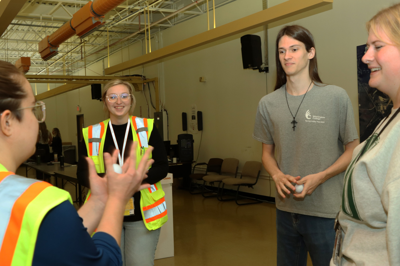 Four individuals stand in a room and talk to each other, two of the individuals wear yellow safety vests