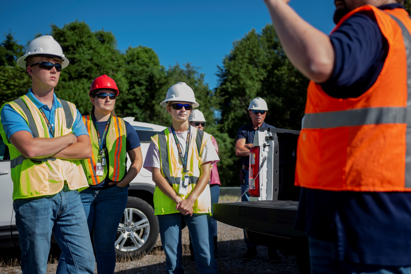 Three student interns stand in the foreground in hardhats and safety vests and listen to a person with their back facing the camera giving a tour
