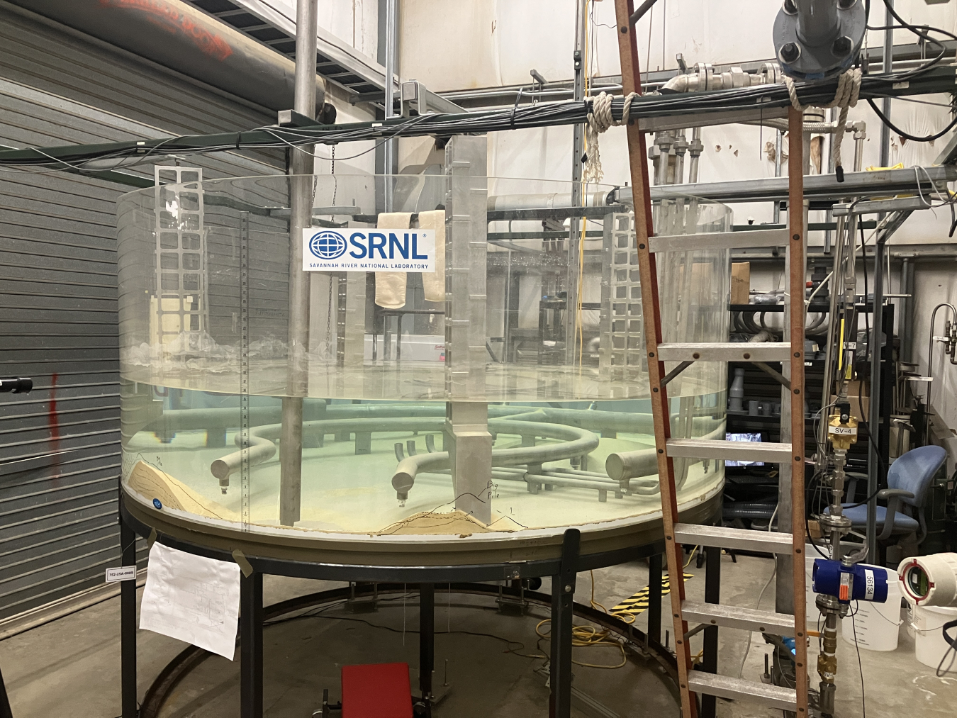 A large, round, glass table with clear liquid inside, it has pipes running through it and a ladder behind it