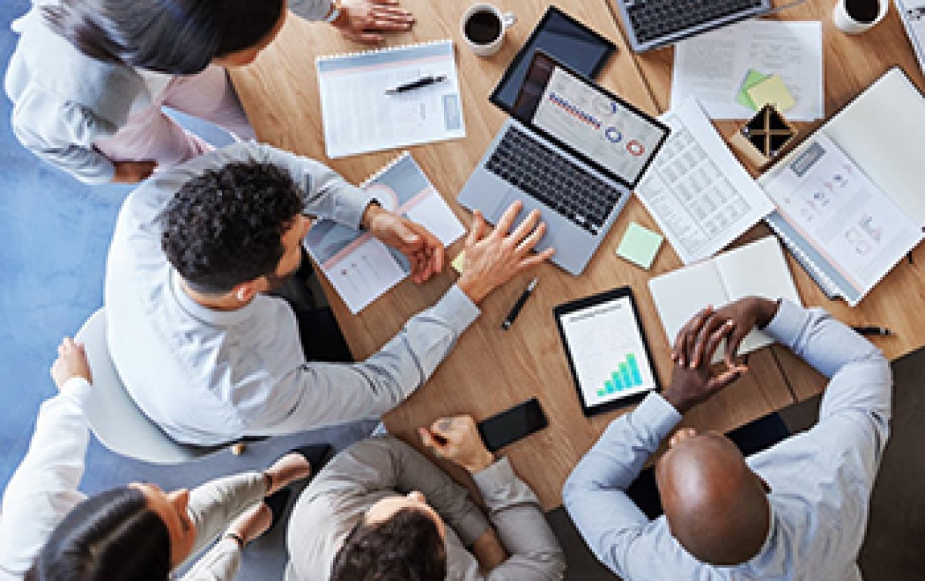 Photo looking down at a conference table surrounded by five people. On the table are laptops, tablets, and notebooks.