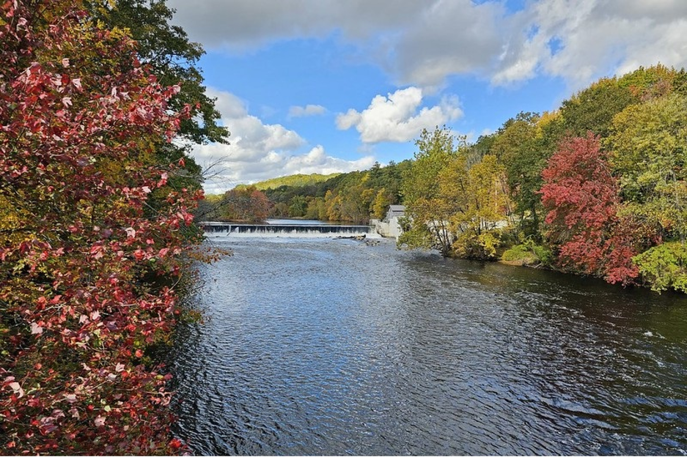 Downriver view of Albion Dam in Cumberland, Rhode Island, with red and green foliage on trees on the river banks.