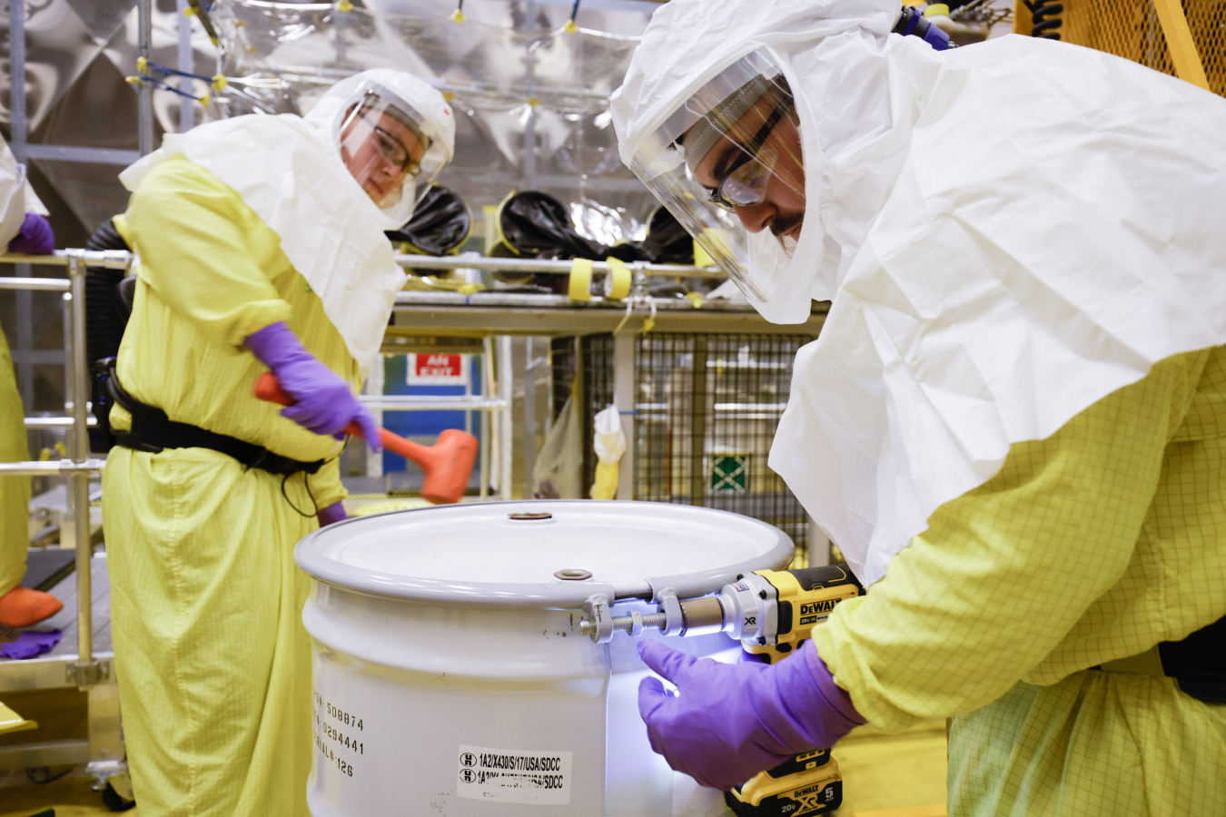 Two employees in yellow and white hazmat suits hold drills and hammers and work on a storage drum