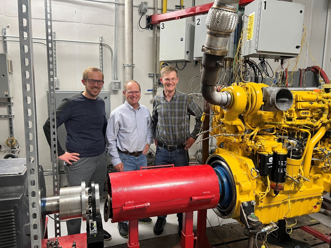 Three men stand behind a large piece of red and yellow equipment in a lab setting.
