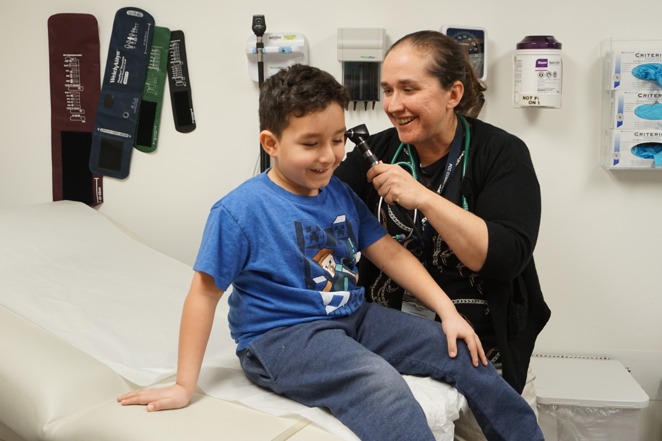 A health care provider looks into the ear of a child sitting on an exam table in an examination room.