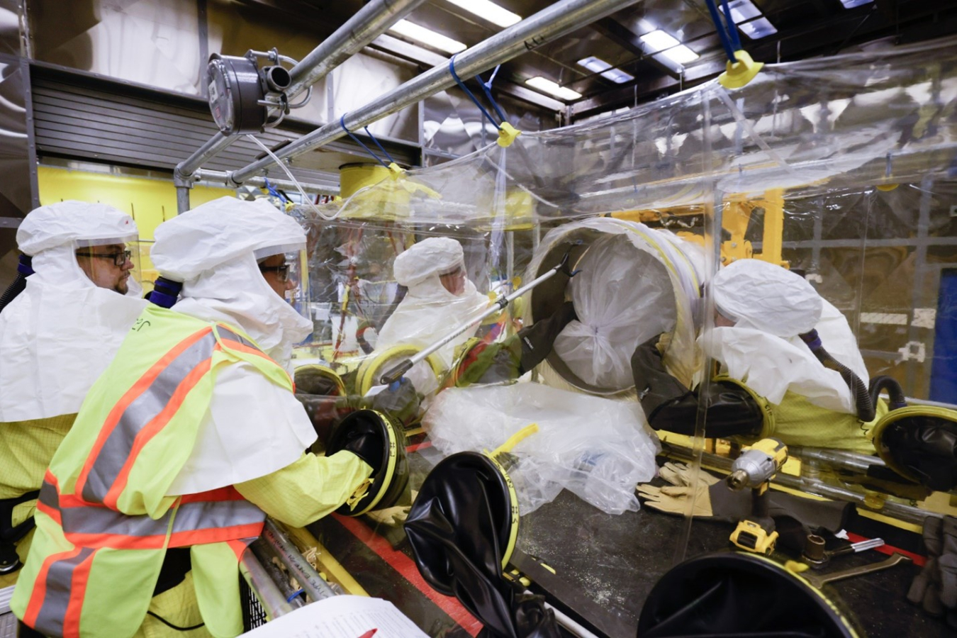 A group of employees in yellow and white hazmat suits using a glovebox machine