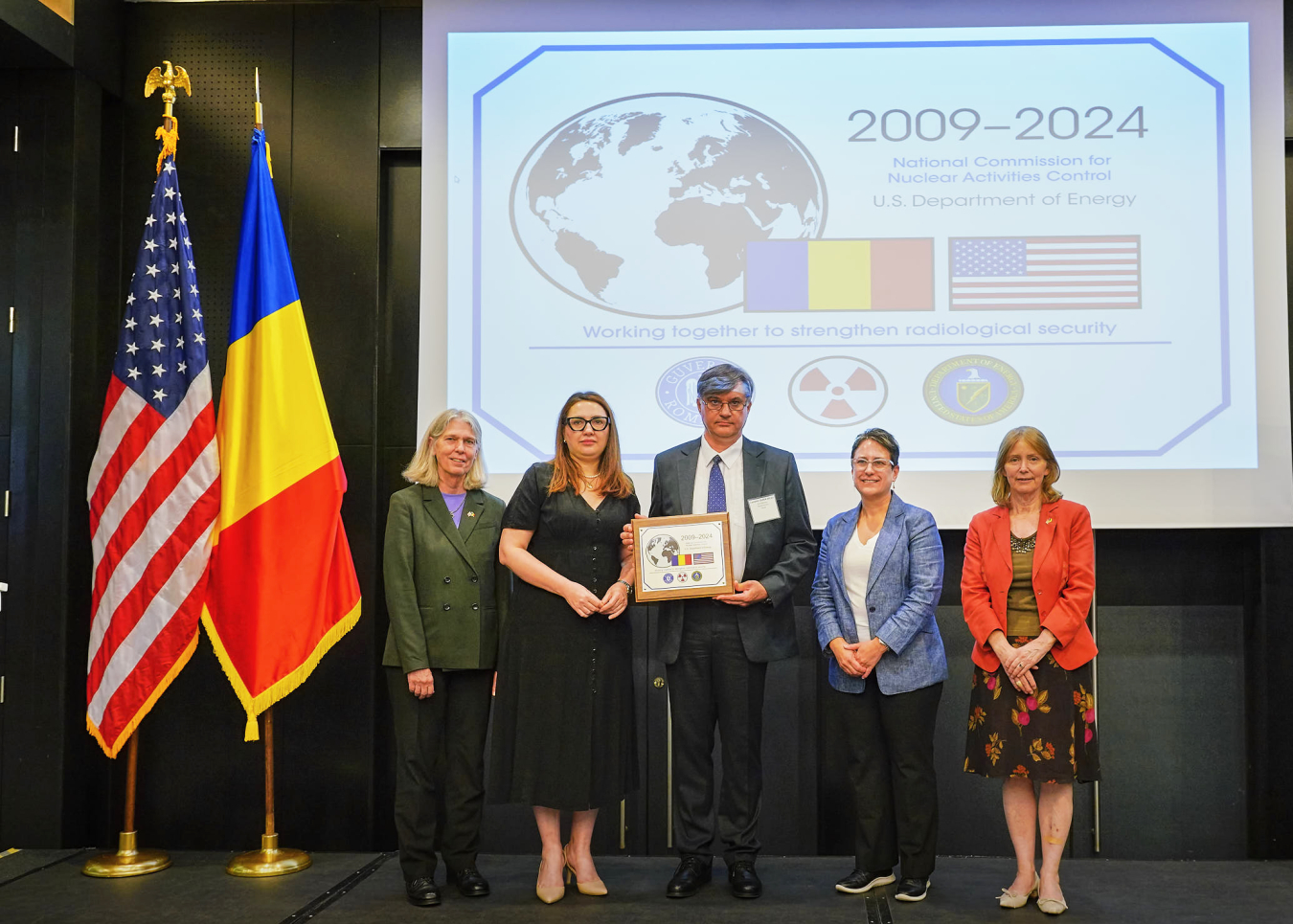 A group of five people stand next to U.S. and Romanian flags. Behind them is a slide on the wall that says 2009-2024 and the names of the two organizations.
