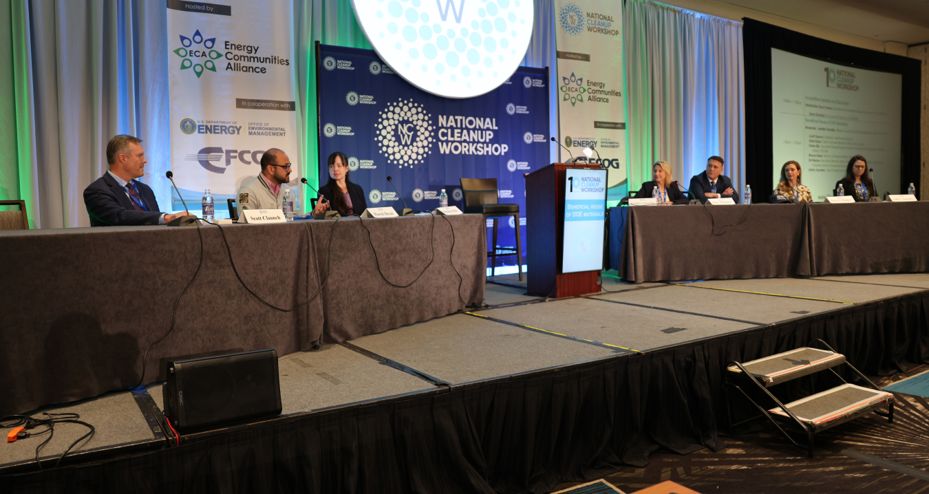 Department of Energy leaders sit at tables on either side of a stage and carry out a panel at a conference
