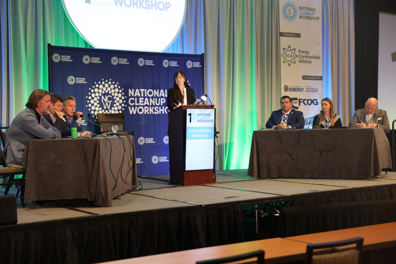 DOE leaders sit on either side of a stage at a table with another standing behind a podium in the middle of the stage