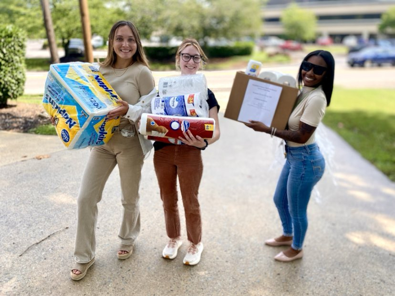 Three women stand on a sidewalk outside and hold toilet paper and paper towels