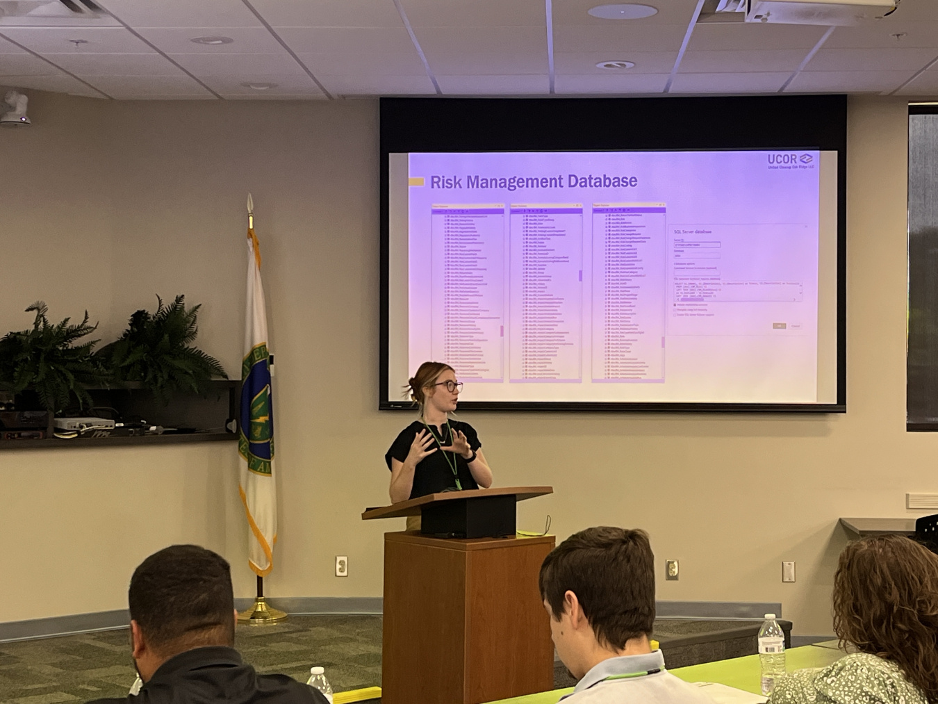 A woman standing at the front of a room behind a podium gives a presentation to people sitting at tables in the room