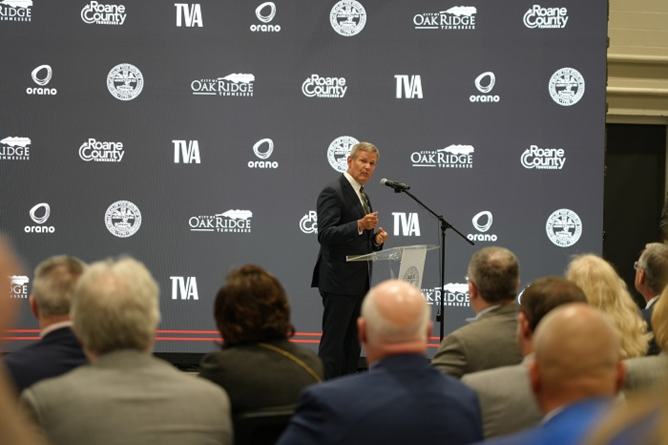 A man stands behind a podium on stage and speaks into a microphone while an audience watches him