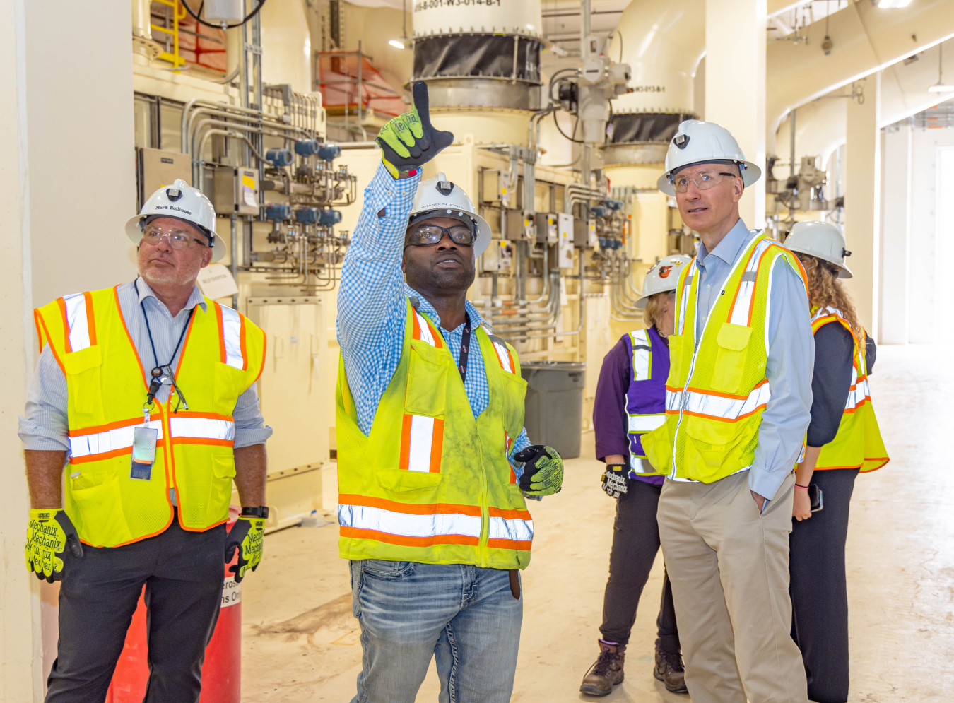 DOE leaders and site workers tour a facility, they wear white hard hats and yellow safety vests