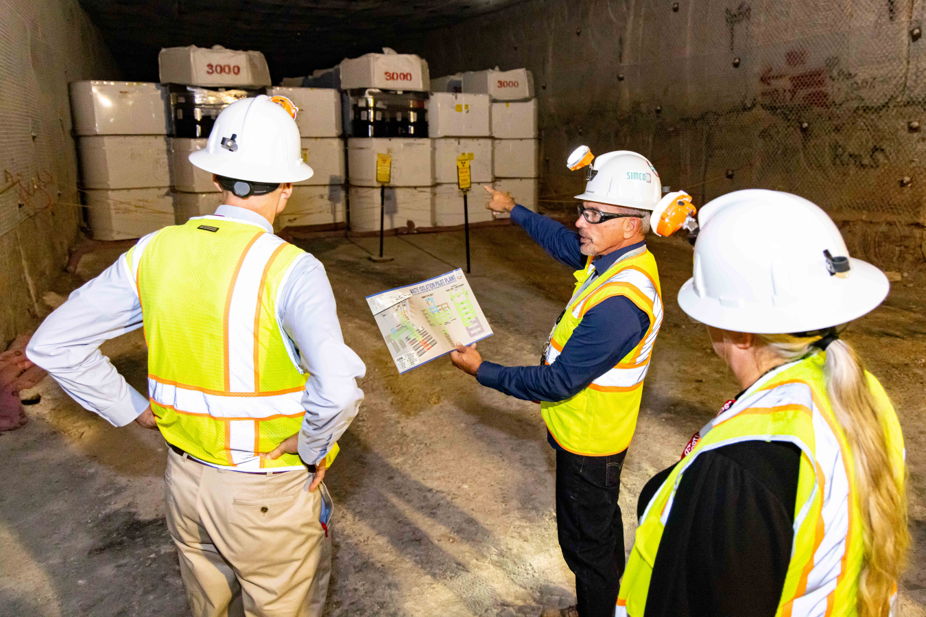 A man conducts a tour and points to an object while speaking to two others, they wear white hard hats and yellow safety vests