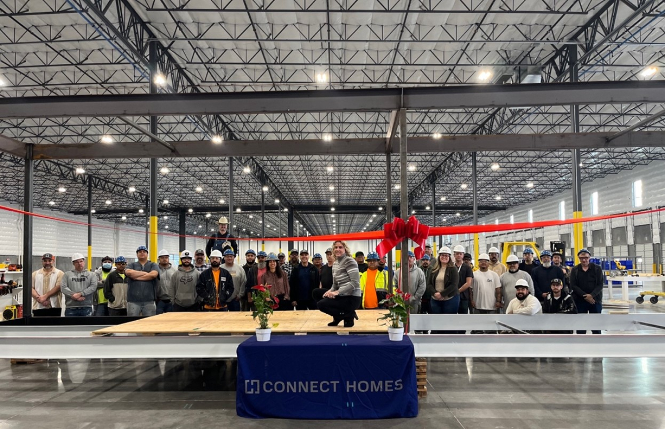 The view of the inside of a large manufacturing plant with high ceilings and a large group of workers facing the camera and smiling, with steel girders above them.