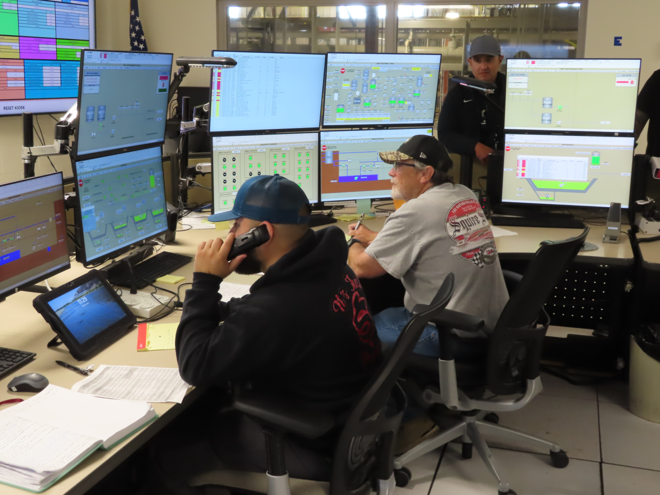 Two employees sit at desks with computers and look at the screens, one employee is also on the phone