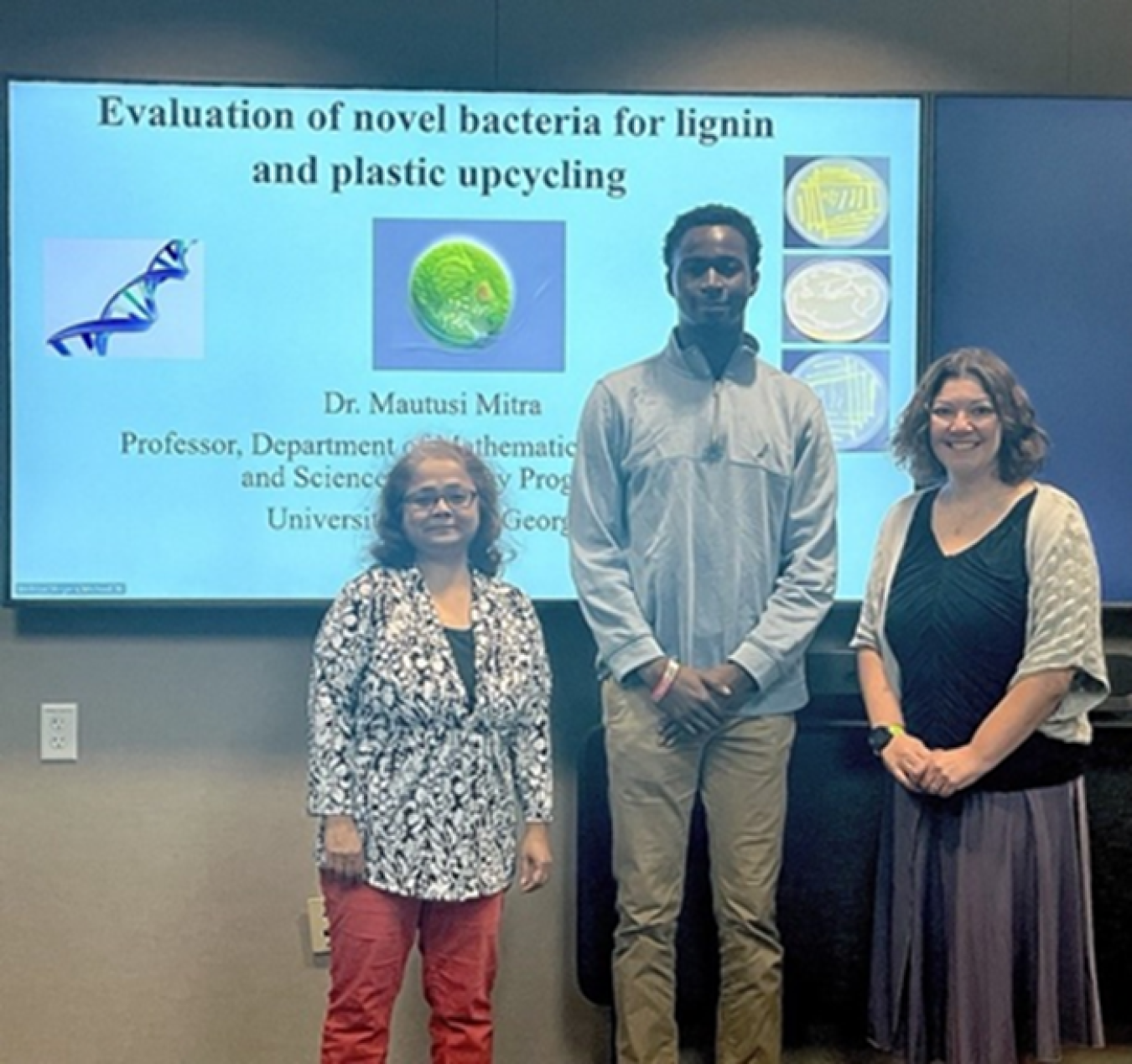 Three people are standing in front of a large slide that says, “Evaluation of novel bacteria for lignin and plastic upcycling”. The slide is blue with a DNA strand and pictures of bacteria. From left to right the picture shows University of West Georgia Professor Mautusi Mitra (left) and student Chukwuemeka Okpala (center) and Oak Ridge National Laboratory scientist Carrie Eckert on the right. The group presented their research findings on plastic upcycling during the DOE Visiting Faculty Program.