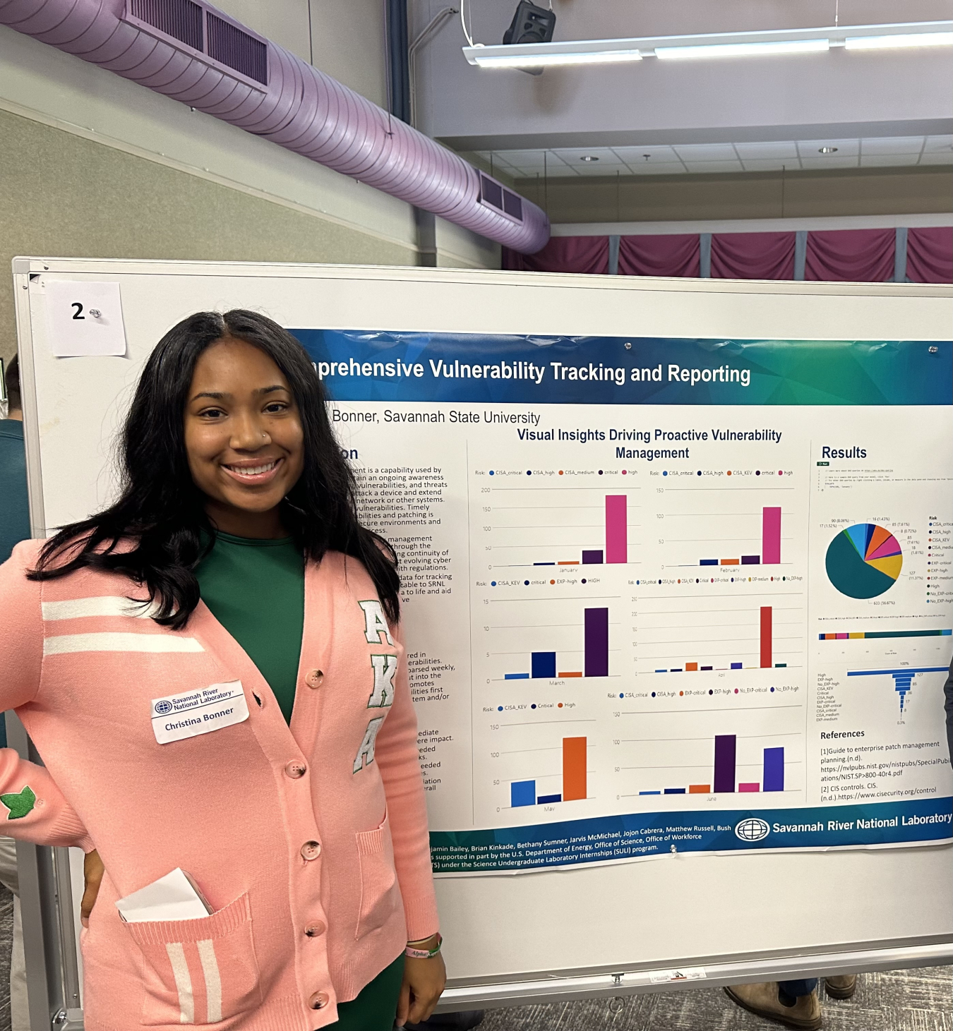 Christina Bonner, a black woman with a wide smile and shoulder-length black hair, is standing in front of a poster entitled “Comprehensive Vulnerability Tracking and Reporting”. She is wearing a peach-colored sweater and green shirt. The poster is colorful and shows several charts and graphs. In the center of the poster is a subtitle:  “Visual Insights Driving Proactive Vulnerability Management.” The venue is a scientific poster session Bonner attended while a SULI intern. 