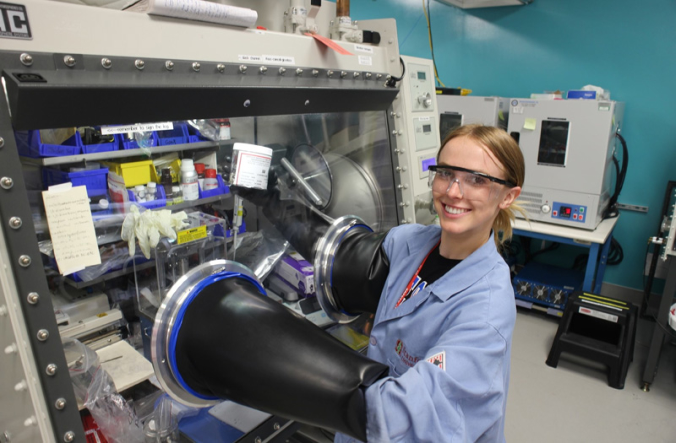 Sarah Cole of Boise State, a student with a wide smile and blond hair that is tied back, is dressed in a blue lab coat and wearing safety glasses while assembling batteries inside an argon glovebox. There are big black tubes where she is able to put her hands through and work within the glovebox at the SLAC-Stanford Battery Center. This method enables researchers to assemble batteries inside an argon glovebox, which stores air-sensitive materials in an oxygen-free environment.