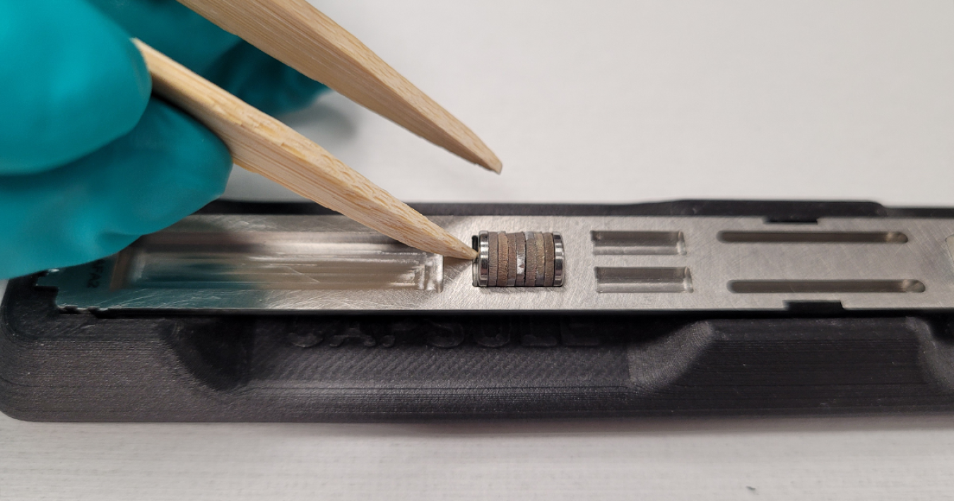 Closeup photo of a gloved hand holding wooden tweezers next to material test capsules