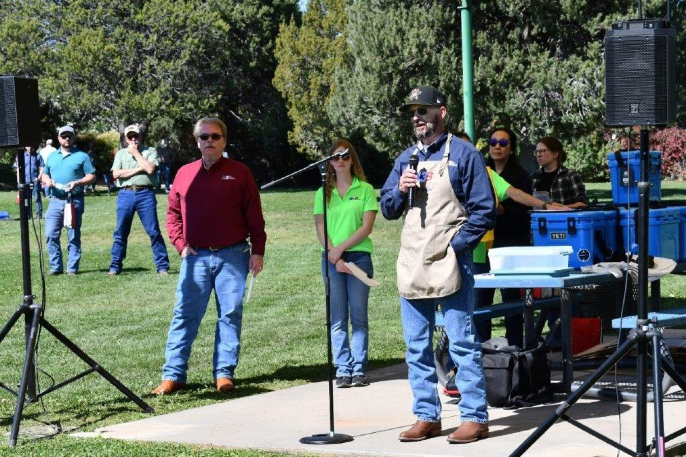 A man stands outside at a safety fair event and holds a microphone and speaks into it. People stand scattered behind him