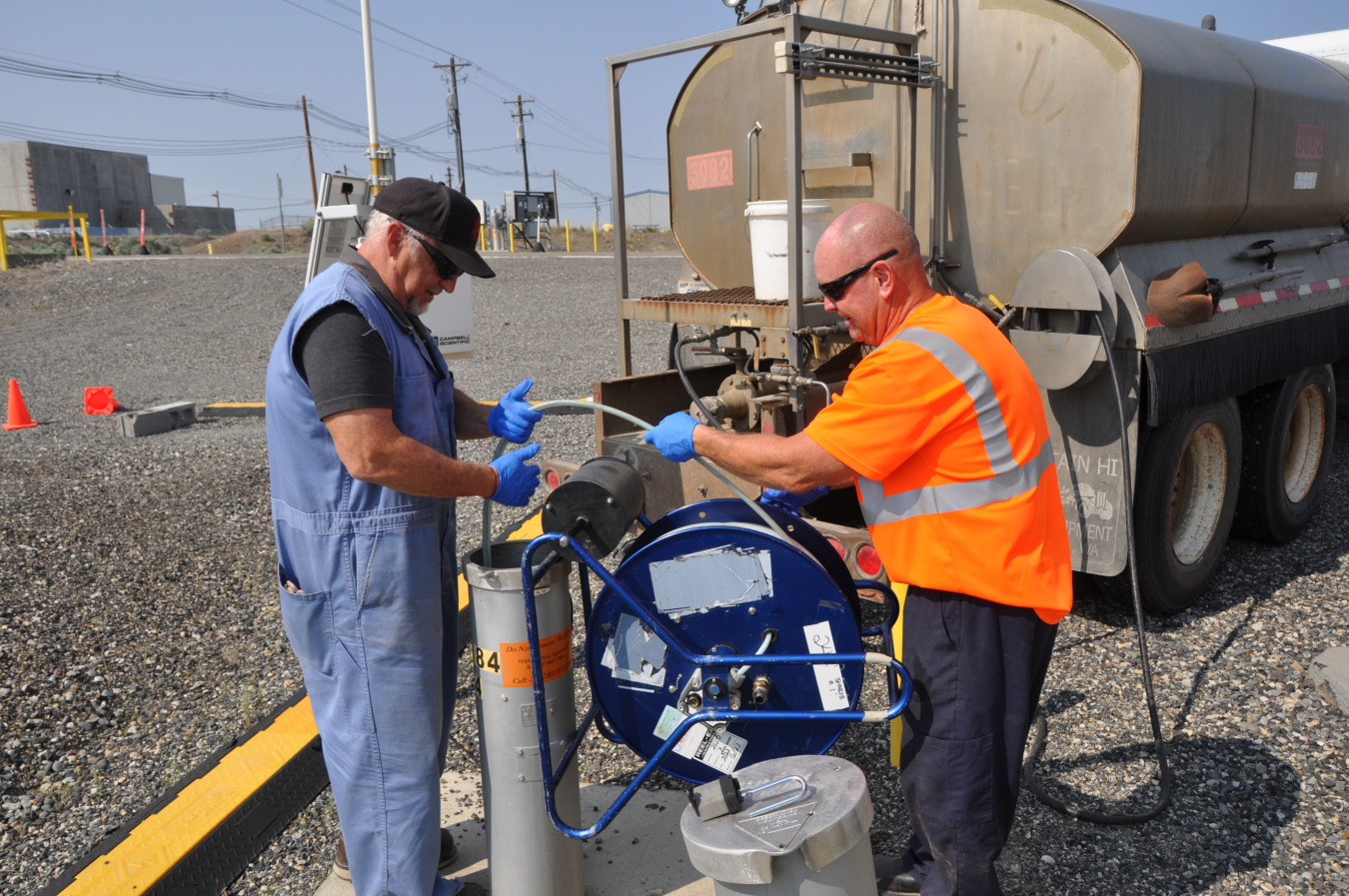 Two men working on a well, they hold a hose and lower it down the well. The hose is attached to a truck 
