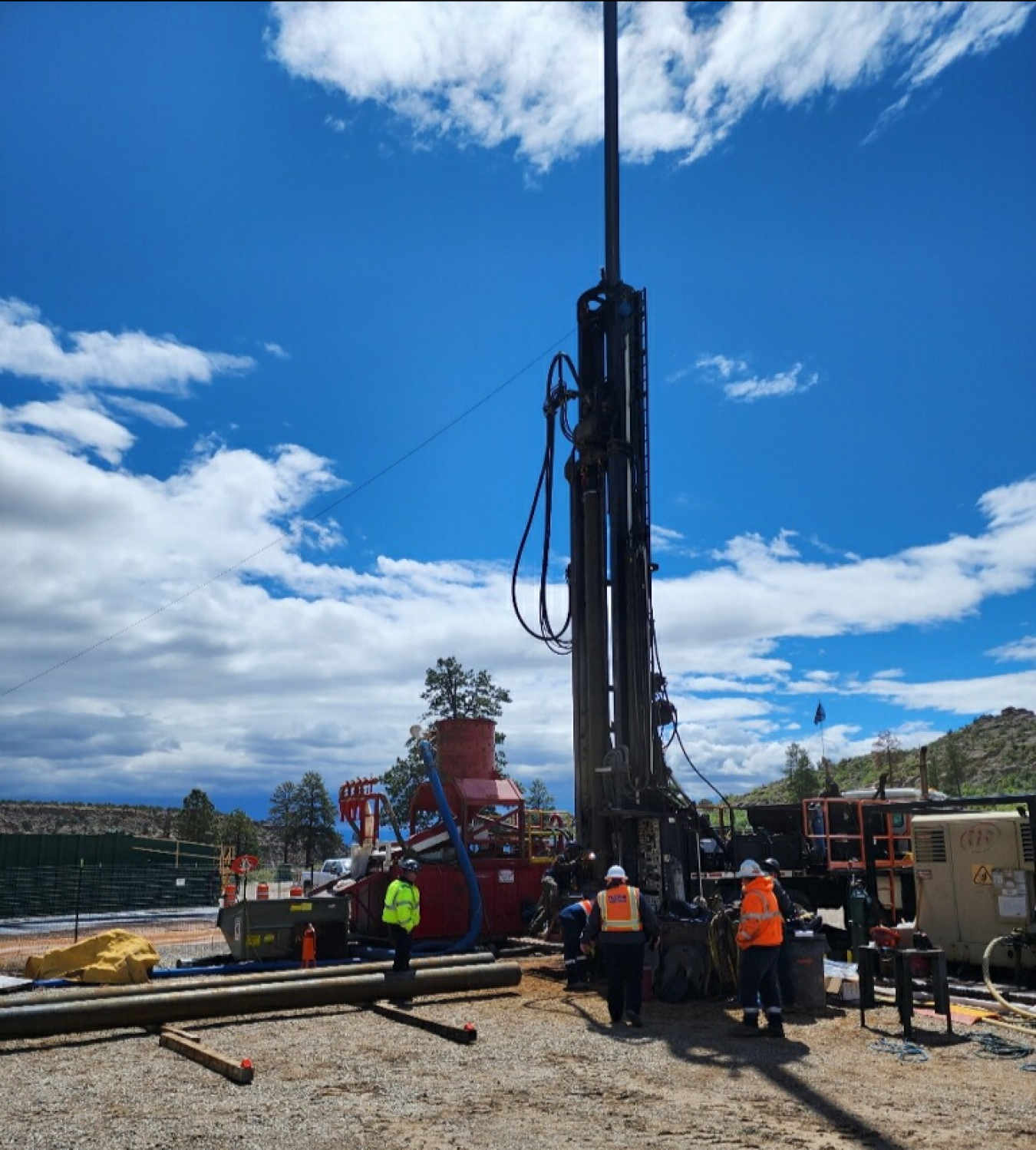 A large drill going into a well in the ground. Employees in neon protective gear stand around and operate it