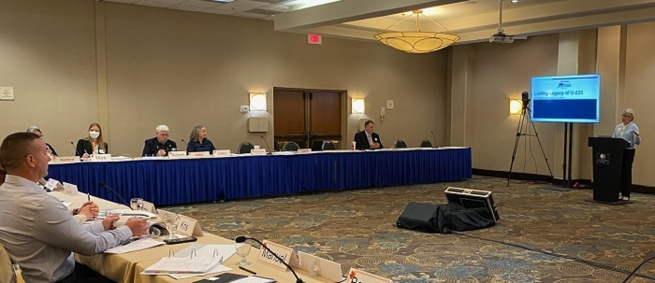 A room with two long tables and people sitting at the tables look at  a man giving a presentation at the front of the room