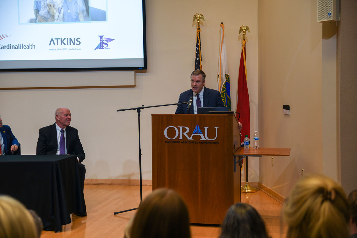 A man stands behind a podium at the front of an auditorium and gives a presentation and addresses the crowd