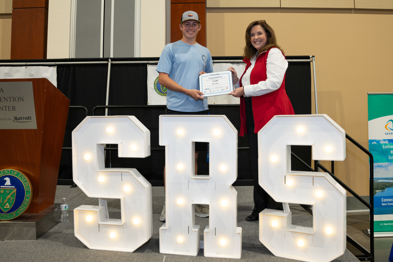 Two people stand behind large white letters that read "SRS"