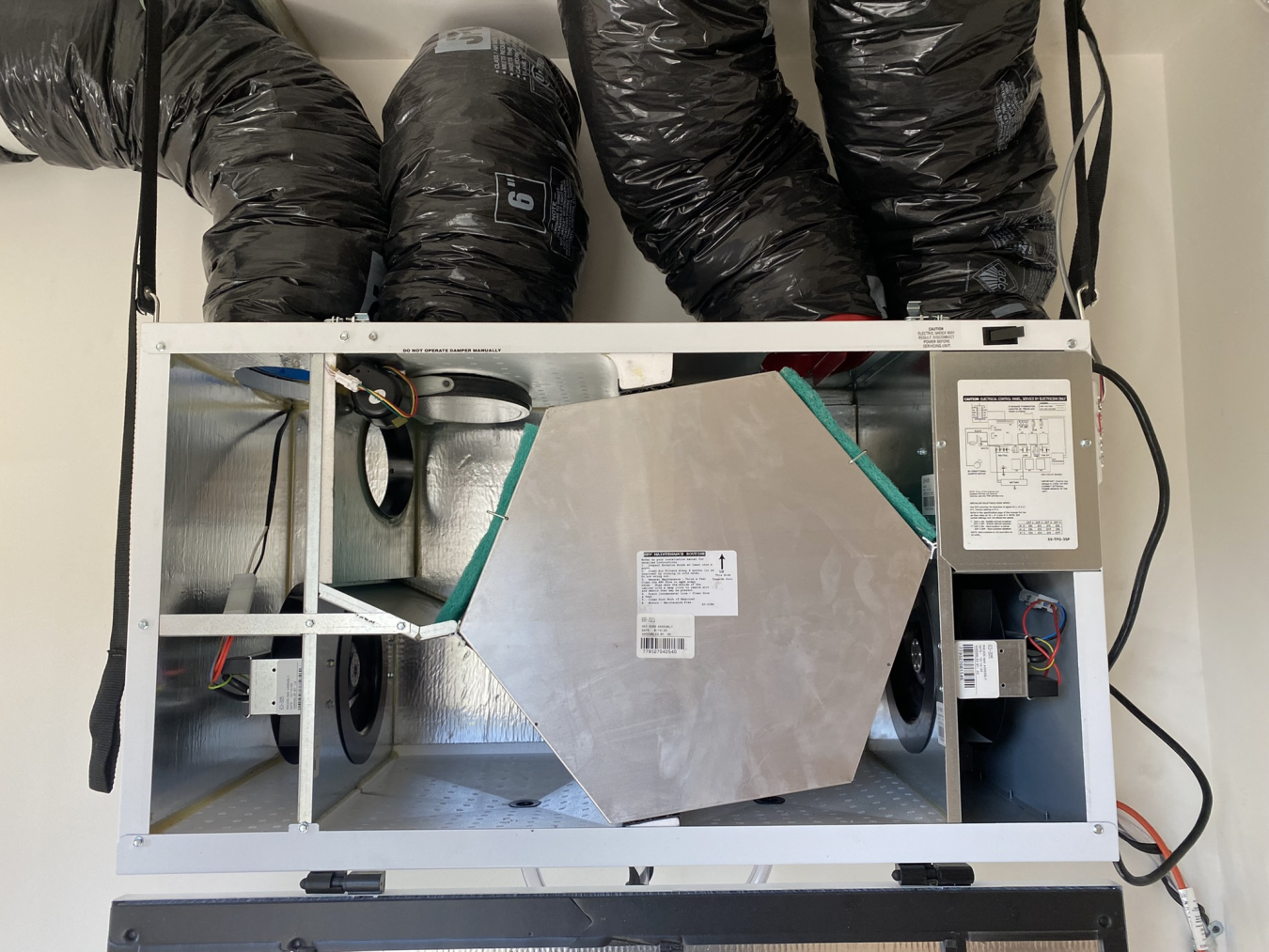 A piece of HVAC equipment mounted to a wall with exposed parts and four ducts connected to the top.